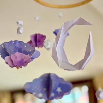 Le mobile Clair de Lune est composé d'un cercle en bambou de 25cm, avec 10 descentes de fil de nylon. Au centre du mobile une lune blanche plié en origami. Autour du cercle les fils sont disposés de sortes à crées une descente progressive de chaque éléments. Sur chaque fil se trouve une étoile blanche en origami, suivi d'un nuage formé à partir de 6 découpe de nuage. Il y a trois tailles de nuage, petit, moyen, grand qui se succèdent. . Les nuages sont mauves avec des motifs léger d'étoiles et de nuages. Ce mobile doux rappellent le début de nuit où les nuages prends une couleur mauve.