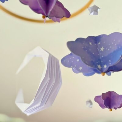 Le mobile Clair de Lune est composé d'un cercle en bambou de 25cm, avec 10 descentes de fil de nylon. Au centre du mobile une lune blanche plié en origami. Autour du cercle les fils sont disposés de sortes à crées une descente progressive de chaque éléments. Sur chaque fil se trouve une étoile blanche en origami, suivi d'un nuage formé à partir de 6 découpe de nuage. Il y a trois tailles de nuage, petit, moyen, grand qui se succèdent. . Les nuages sont mauves avec des motifs léger d'étoiles et de nuages. Ce mobile doux rappellent le début de nuit où les nuages prends une couleur mauve.
