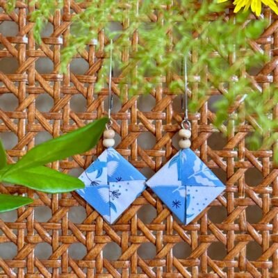 Des boucles d'oreilles avec de longs crochets en acier inoxydable, composées de deux petites perles en bois et d'un pliage en origami de la forme d'un carré plat où deux feuilles s'entremêlent. Le papier est blanc et bleu avec des motifs d'oiseaux et de feuilles de couleur bleu et blanc.
