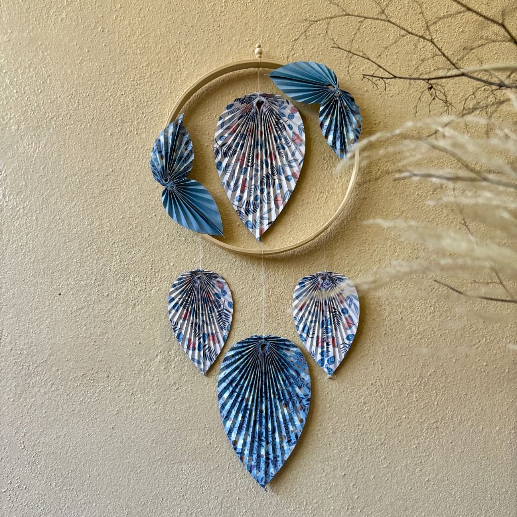 Un cercle en bambou de 15cm, ornée de pliage en origami en forme de feuille, une feuille se trouve dans le centre du cercle, quatre petites feuilles sont collées en diagonale sur le cercle, puis trois feuilles qui pendent le long du cercle. Les feuilles sont de couleurs bleues et blanches avec des motifs fleurit.