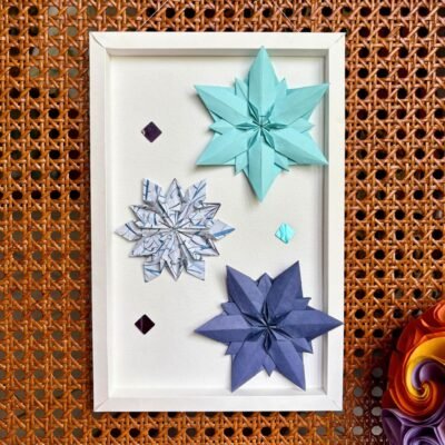 Un cadre rectangle plié à la main, de couleur blanc. A l'intérieur, trois pliages de flocons de neige de couleur bleu clair, bleu foncé, et blanc avec des motifs bleus sont disposé de façon harmonieuse. Trois sequin miroitant les accompagnent.