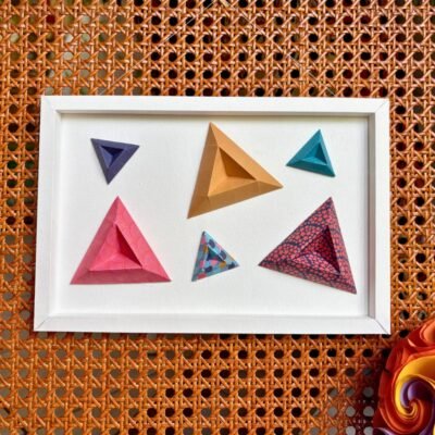 Un cadre rectangle plié à la main, de couleur blanc. A l'intérieur sont disposés six triangles en origami (bleu, dorée, rouge, bleu, bleu et motifs rouge, bleu et motifs rose)