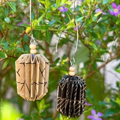 Des boucles d'oreilles avec de longs crochets en acier inoxydable, composées de deux petites perles en bois et d'un pliage en origami qui rappelle la forme des lanternes japonaise. Ces boucles d'oreilles sont en couleurs dépareillées : d'un côté du marron avec des motifs géométriques noirs et de l'autre du noir avec des motifs géométriques marrons.