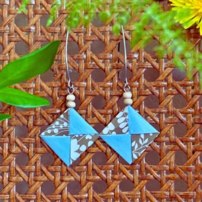 Des boucles d'oreilles avec de longs crochets en acier inoxydable, composées de deux petites perles en bois et d'un pliage en origami de la forme d'un carré plat où deux feuilles s'entremêlent. L'un des papiers est bleu sans motifs et l'autre est dorée avec des motifs de feuilles blanc.