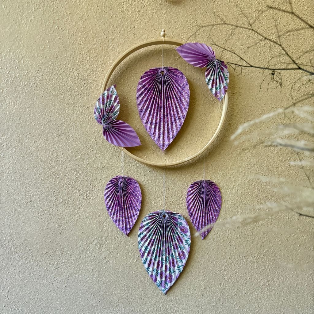 Un cercle en bambou de 15cm, ornée de pliage en origami en forme de feuille, une feuille se trouve dans le centre du cercle, quatre petites feuilles sont collées en diagonale sur le cercle, puis trois feuilles qui pendent le long du cercle. Les feuilles sont de couleurs mauves et blanches avec des motifs fleurit.