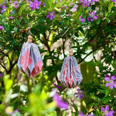 Des boucles d'oreilles avec des crochets simples en acier inoxydable, composées de deux petites perles en bois et d'un pliage en origami qui rappelle la forme d'une goutte d'eau. Ces boucles d'oreilles sont blanc, gris et orange avec divers motifs fleurit et géométrique.