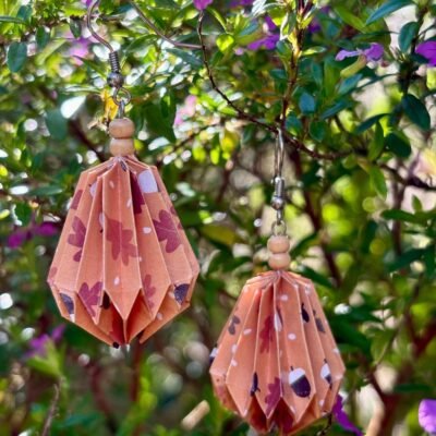 Des boucles d'oreilles avec des crochets simples en acier inoxydable, composées de deux petites perles en bois et d'un pliage en origami qui rappelle la forme d'une goutte d'eau. Ces boucles d'oreilles sont orange avec des motifs de feuilles et de noisettes marron, blanc.