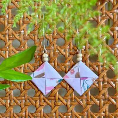 Des boucles d'oreilles avec de longs crochets en acier inoxydable, composées de deux petites perles en bois et d'un pliage en origami de la forme d'un carré plat où deux feuilles s'entremêlent. Le papier est rose avec de léger motifs fleurit.