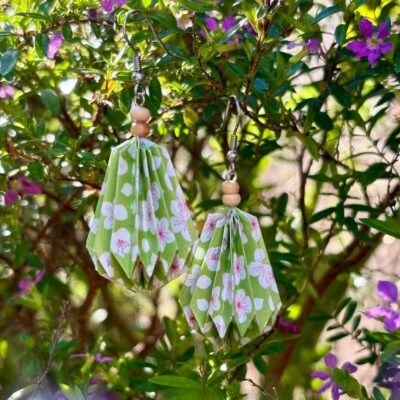 Des boucles d'oreilles avec des crochets simples en acier inoxydable, composées de deux petites perles en bois et d'un pliage en origami qui rappelle la forme d'une goutte d'eau. Ces boucles d'oreilles sont vertes avec des motifs de fleurs blanches.