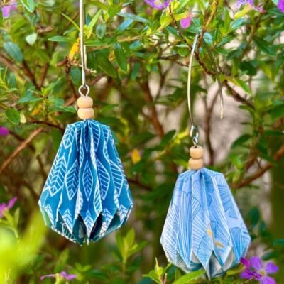 Des boucles d'oreilles avec de longs crochets en acier inoxydable, composées de deux petites perles en bois et d'un pliage en origami qui rappelle la forme d'une goutte d'eau près à tomber de sa feuille. Ces boucles d'oreilles sont en couleurs dépareillées : d'un côté du bleu avec des motifs blancs en forme de feuilles, de l'autre du blanc avec des motifs des feuilles gingko en bleus, et des libellules en dorée.