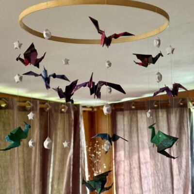 Le mobile dragon est composé d'un cercle en bambou de 25cm, avec 10 descentes de fil de nylon. Le papier utilisé pour les dragons est dans un style galaxie de différente couleur, il est également composé d'étoile en origami appelés "étoile de la chance" de couleur blanche. Au centre du mobile se trouve un gros dragon mauve en dessous de lui un dragon plus petit de couleur verte. Les 9 descentes autour du cercle se déclinent de cette façon : Descente 1 = 1 dragon en haut puis 2 étoiles ; descente 2= 1 étoile, 1 dragon, 1 étoile ; descente 3 = 2 étoiles 1 dragon. Ce modèle est répété sur les 6 autres descentes de façon harmonieuse. Les dragons sont de couleur bleu/verte, rouge/mauve, mauve/bleu. Ce mobile invite à la rêverie, à l'aventure, avec ces dragons majestueux voletant légèrement.