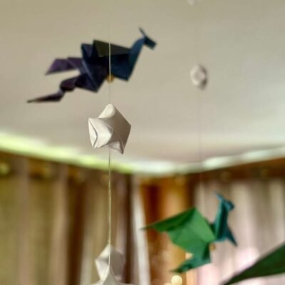 Le mobile dragon est composé d'un cercle en bambou de 25cm, avec 10 descentes de fil de nylon. Le papier utilisé pour les dragons est dans un style galaxie de différente couleur, il est également composé d'étoile en origami appelés "étoile de la chance" de couleur blanche. Au centre du mobile se trouve un gros dragon mauve en dessous de lui un dragon plus petit de couleur verte. Les 9 descentes autour du cercle se déclinent de cette façon : Descente 1 = 1 dragon en haut puis 2 étoiles ; descente 2= 1 étoile, 1 dragon, 1 étoile ; descente 3 = 2 étoiles 1 dragon. Ce modèle est répété sur les 6 autres descentes de façon harmonieuse. Les dragons sont de couleur bleu/verte, rouge/mauve, mauve/bleu. Ce mobile invite à la rêverie, à l'aventure, avec ces dragons majestueux voletant légèrement.