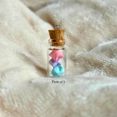 La fiole n°1 est une fiole en verre d'environs 3cm en hauteur et est refermé par un bouchon en liège. La fiole est rempli par 3 minuscule étoiles coloré de rose, mauve et bleu. Le bouche en liège contient un petit crochet pour y rajouter un fil au besoin.