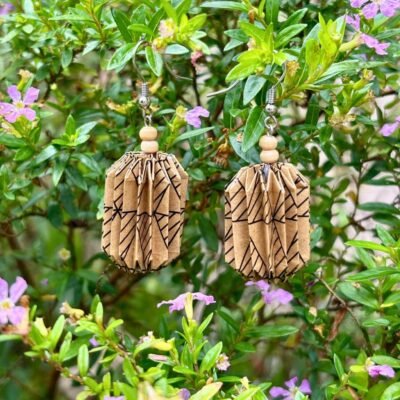 Des boucles d'oreilles avec de longs crochets en acier inoxydable, composées de deux petites perles en bois et d'un pliage en origami qui rappelle la forme des lanternes japonaise. Ces boucles d'oreilles sont marrons avec des motifs géométriques noirs.