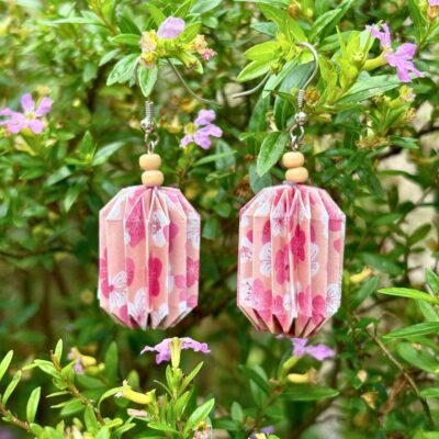 Des boucles d'oreilles avec de longs crochets en acier inoxydable, composées de deux petites perles en bois et d'un pliage en origami qui rappelle la forme des lanternes japonaise. Ces boucles d'oreilles sont beiges avec des motifs de fleurs roses.