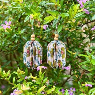 Des boucles d'oreilles avec de longs crochets en acier inoxydable, composées de deux petites perles en bois et d'un pliage en origami qui rappelle la forme des lanternes japonaise. Ces boucles d'oreilles sont beiges avec des motifs de fleurs vertes et de feuilles bleues.