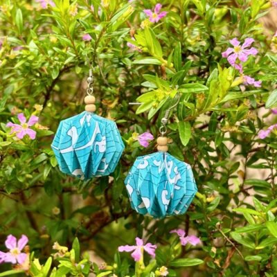 Des boucles d'oreilles avec des crochets simples en acier inoxydable, composées de deux petites perles en bois et d'un pliage en origami qui rappelle la forme des lanternes japonaise ronde ou d'une perle. Ces boucles d'oreilles sont bleues avec des motifs de vagues blanches.