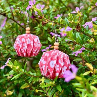 Des boucles d'oreilles avec des crochets simples en acier inoxydable, composées de deux petites perles en bois et d'un pliage en origami qui rappelle la forme des lanternes japonaise ronde ou d'une perle. Ces boucles d'oreilles sont roses avec des motifs de fleurs blanches.
