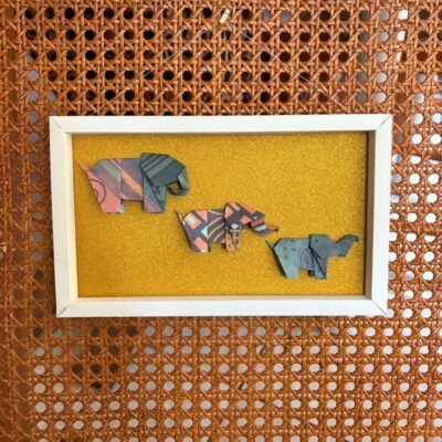 Un cadre rectangle plié à la main, de couleur blanc et dorée brillant. A l'intérieur, trois éléphants en origami y sont disposé, et de taille dégressive.